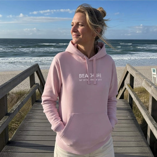 Blonde Frau trägt einen rosa Sylt Hoodie mit einem weißen Schriftzug Beachlife und den Koordinaten von Sylt. Die Frau steht an einem Strandaufgang. Im Hintergrund sieht man die Nordsee.